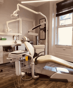 Voyagerili dental chair with overhead light in bright dental clinic room, brushed wood flooring, window with blinds, and framed certificates on wall for Hewett's Dental services in Brighouse, Huddersfield, Halifax, Bradford.